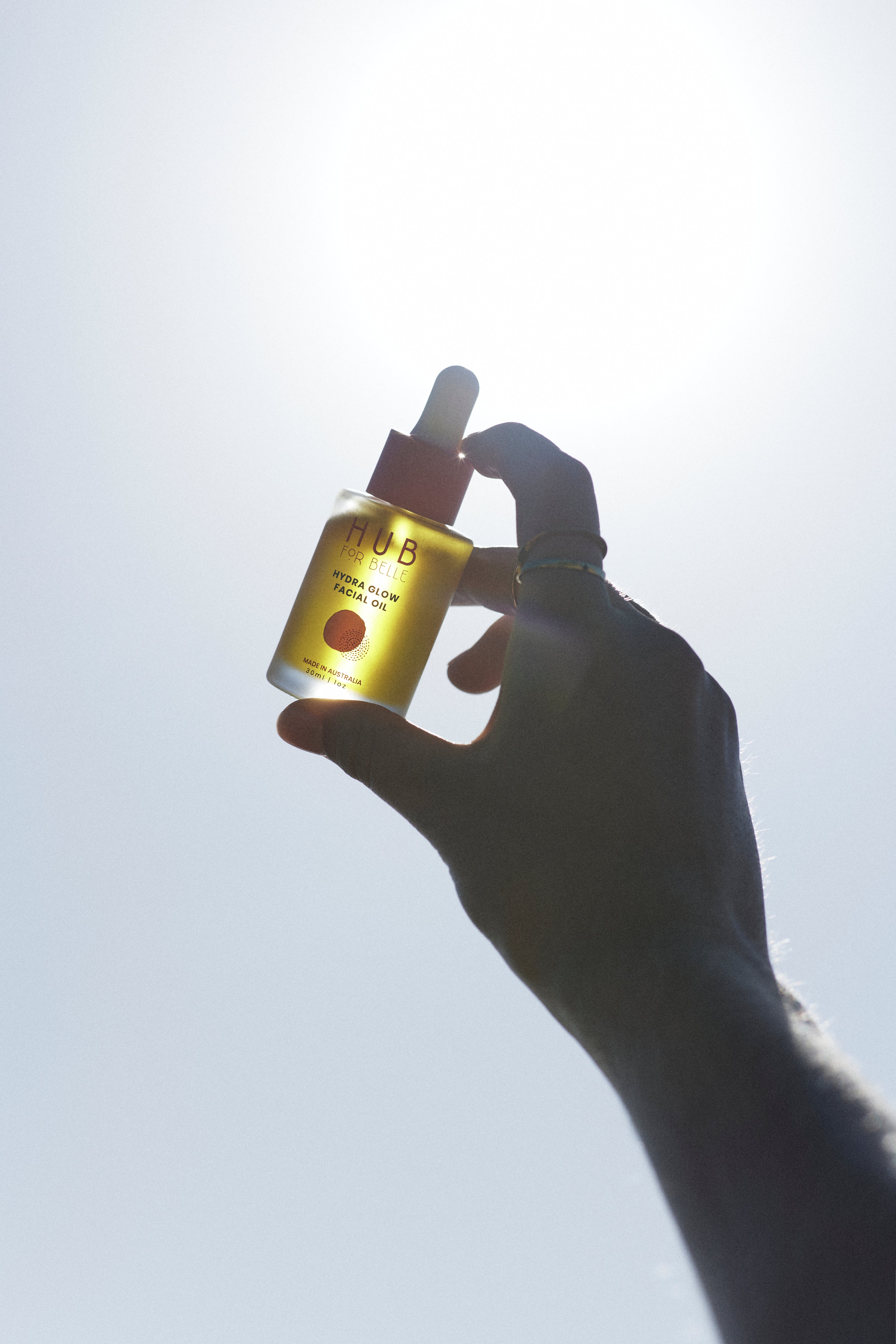 Hand holding to sun a bottle of Hydra Glow Facial oil made with natural vegan Australian botanicals for glowing skin from Hub for Belle