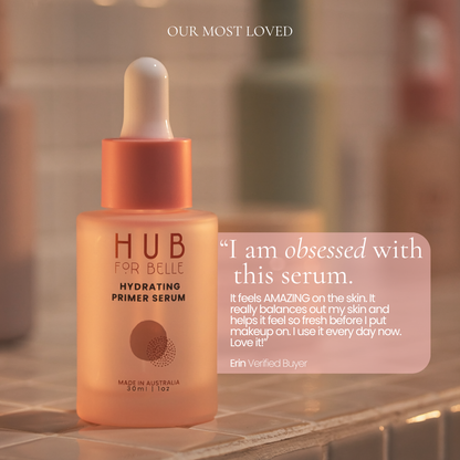 A bottle of HUB FOR BELLE Hydrating Primer Serum in a warm peach-orange color with a white dropper cap, set on a tiled bathroom counter with a soft-focus background. A pink text box on the right features a customer testimonial.